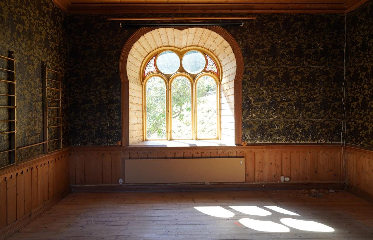 Et tomt rom i villaen på Lysøen, med et stort vindu midt på veggen, hvor lyset faller inn. Rikt ornamentert tapet på veggene.