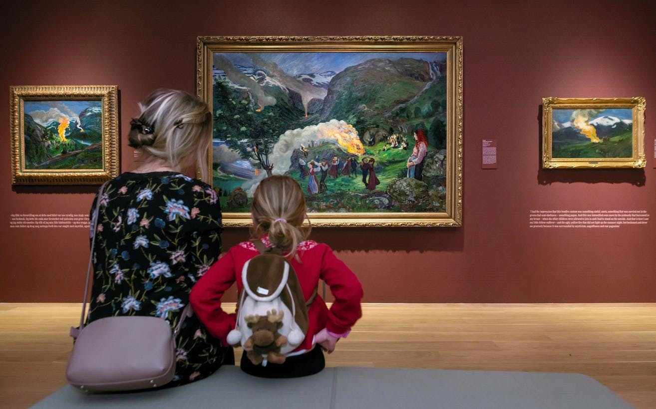 En kvinne og et barn sitter i en utstilling, foran malerier av Nikolai Astrup